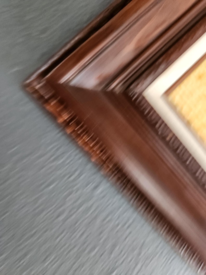 May include: Close-up of a dark brown wooden picture frame with a textured surface. The frame has a beveled edge and a cream-colored matting around the artwork. The background is a blurred gray.