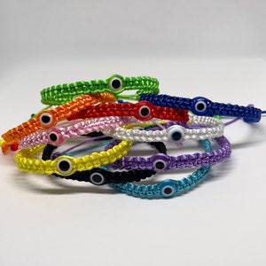 May include: A stack of colourful bracelets with evil eye charms. The bracelets are made of woven string and come in a variety of colours, including green, red, pink, white, yellow, purple, black, and blue.