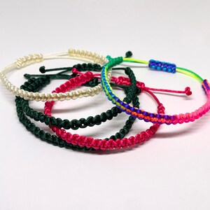 May include: Five colorful friendship bracelets made of braided string. The bracelets are in shades of white, green, pink, and rainbow.