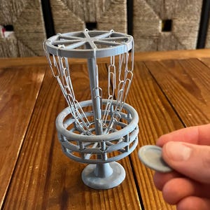 May include: A miniature gray disc golf basket with a chain net, set on a wooden surface. A gray disc is held nearby. The basket has a circular base and a series of metal chains hanging from the top.