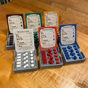 May include: A board game setup with various colored cards labeled "BUILDING COSTS" and miniature building pieces. The game pieces are in gray trays, with white, red, green, blue, and brown building components.