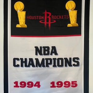 Könnte beinhalten: Ein weißes und schwarzes Banner mit dem Houston Rockets Logo und dem Text "NBA CHAMPIONS 1994 1995". Das Banner hat einen roten und schwarzen Rand und acht weiße Sterne.