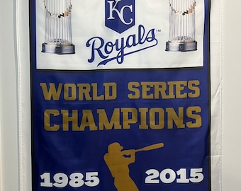 May include: A blue and white flag with the Kansas City Royals logo and the text "World Series Champions 1985 2015". The flag features two silver trophies and a silhouette of a baseball player.
