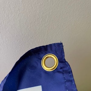 Puede incluir: Primer plano de una bandera azul con un cuadrado blanco y un rect&aacute;ngulo naranja. La bandera tiene un ojal de lat&oacute;n en la esquina superior.