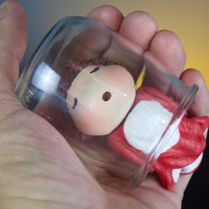 Puede incluir: Un tarro de cristal transparente que contiene una pequeña figura. La figura tiene una cara pálida con ojos negros y un atuendo rojo y blanco. El tarro se sostiene en una mano, con un fondo amarillo y negro.