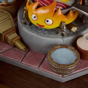 以下が含まれることがあります： 笑顔の顔をした燃えるようなキャラクター、ミニチュアのフィギュア、水で満たされた木製の浴槽が特徴の、詳細なフィギュア。 ふいごと小さな水差しも表示されます。 ベースは石のような表面です。