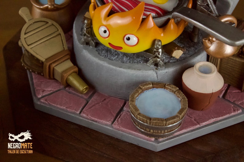 以下が含まれることがあります： 笑顔の顔をした燃えるようなキャラクター、ミニチュアのフィギュア、水で満たされた木製の浴槽が特徴の、詳細なフィギュア。 ふいごと小さな水差しも表示されます。 ベースは石のような表面です。