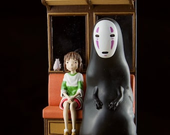 以下が含まれることがあります： アニメ映画のキャラクターを描いた精巧なフィギュア。緑と白のストライプのシャツを着た女の子と、白いマスクをした黒い姿が描かれています。舞台はオレンジ色の座席と茶色のフレームの電車です。