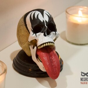 Könnte beinhalten: Ein weißer Schädel mit einem schwarz-weiß bemalten Gesicht, das einem Rockmusiker ähnelt, mit einer roten Zunge, die herausragt. Der Schädel steht auf einem schwarzen Sockel.