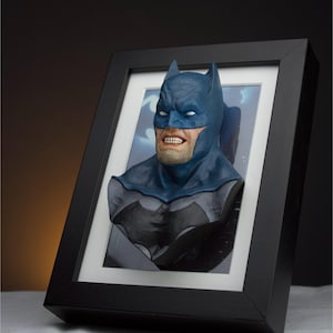 Könnte beinhalten: Eine gerahmte 3D-Büste von Batman mit einem dunkelblauen und grauen Kostüm. Die Büste ist vor einem schwarzen Hintergrund platziert und in einem schwarzen Rahmen mit einem weißen Passepartout gerahmt.