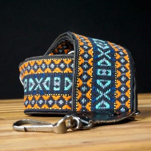 May include: A guitar strap featuring a woven fabric pattern in black, orange, and light blue. The strap has a dark leather end piece with a metal swivel hook attachment. The design consists of repeating geometric shapes and diamond motifs.