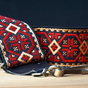 May include: A black guitar strap featuring a repeating geometric pattern in red and tan thread. The strap includes dark leather end tabs and a silver-toned metal swivel hook attachment for securing the instrument to the strap securely.
