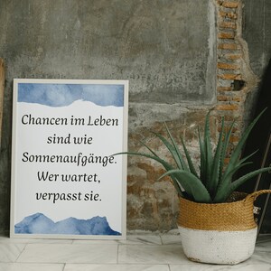 May include: A framed print with the text "Chancen im Leben sind wie Sonnenaufgänge. Wer wartet, verpasst sie." in black font, with blue watercolor accents. A potted aloe vera plant in a woven basket and a wooden crate are also in the image.