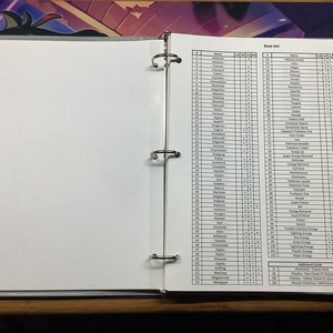 May include: A black and white binder with a list of Pokemon cards and their corresponding numbers. The binder has a white label that says "Base Set."