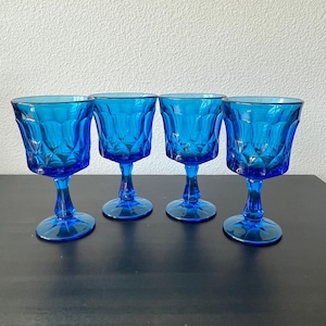 Può includere: Quattro calici blu cobalto con un motivo a diamante. I calici sono disposti in fila su una superficie scura, con un muro chiaro sullo sfondo. I calici hanno un design classico e vintage.