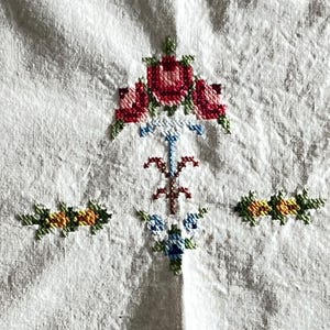 May include: Close-up of a white fabric with cross-stitch embroidery. The embroidery features a central floral design with red roses and green leaves, and smaller floral accents in yellow, orange, and blue.