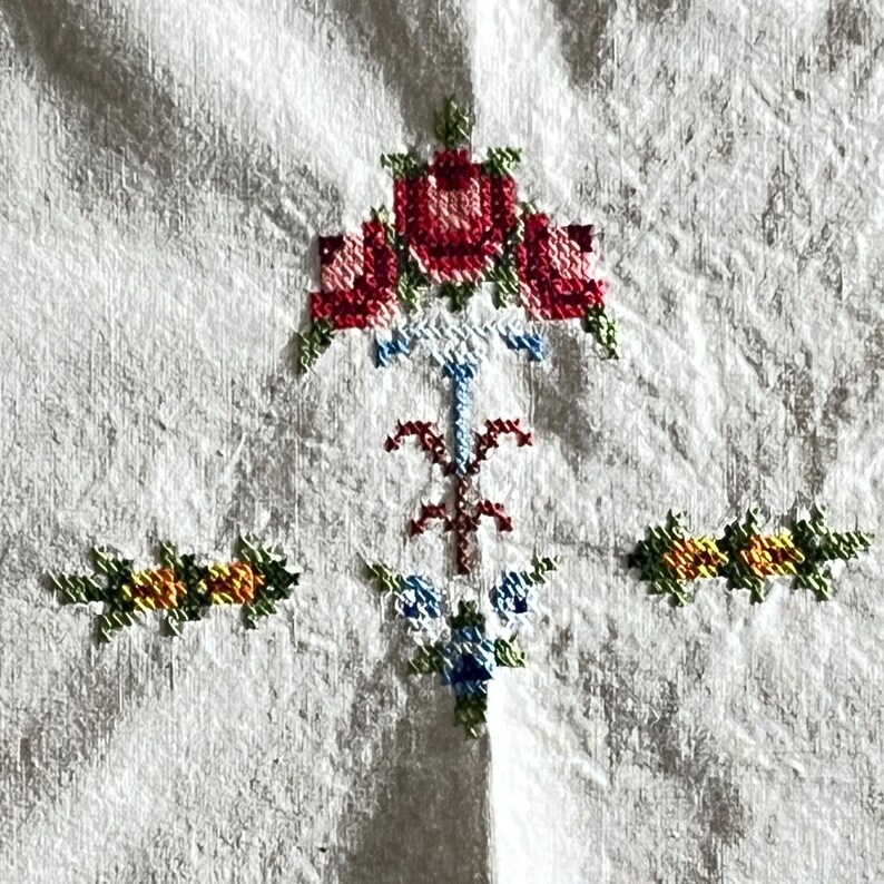 May include: Close-up of a white fabric with cross-stitch embroidery. The embroidery features a central floral design with red roses and green leaves, and smaller floral accents in yellow, orange, and blue.