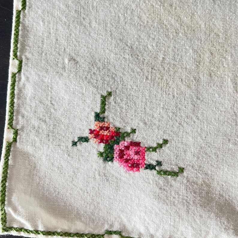 May include: A square, off-white linen cloth with a cross-stitched floral design in the corner. The flowers are pink and red with green stems and leaves. The edges are trimmed with green stitching.