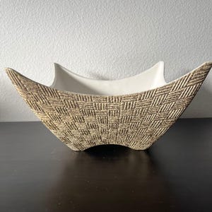 May include: A ceramic bowl with a four-pointed star shape and a smooth white interior. The exterior features a textured, tan and brown geometric pattern consisting of small, repeating rectangular blocks arranged in a woven, basket-like design.