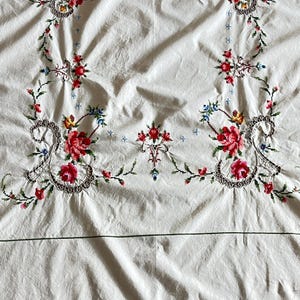 May include: White tablecloth with intricate floral embroidery. The embroidery features red, pink, yellow, and blue flowers with green leaves and stems. The design is symmetrical, with floral patterns at the corners and center.