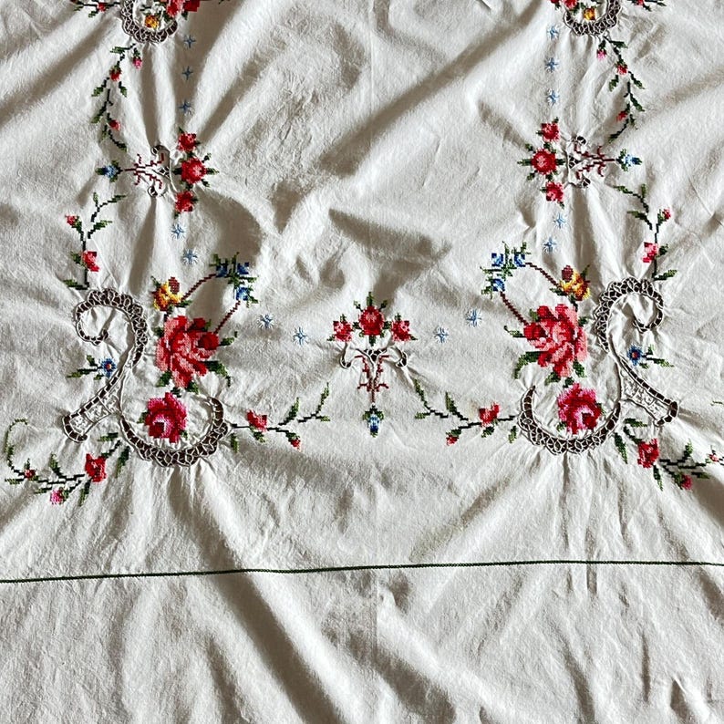 May include: White tablecloth with intricate floral embroidery. The embroidery features red, pink, yellow, and blue flowers with green leaves and stems. The design is symmetrical, with floral patterns at the corners and center.