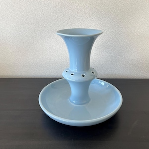 May include: A light blue ceramic flower frog with a flared top and a circular base. The frog has a series of small holes around the middle section for arranging flowers. The base is a shallow dish, and the overall design is simple and elegant.