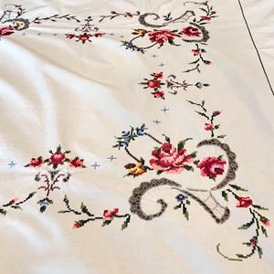 May include: White tablecloth with intricate cross-stitch floral embroidery. The design features red, pink, and blue flowers with green leaves and stems. Decorative gray lace-like accents frame the floral patterns.
