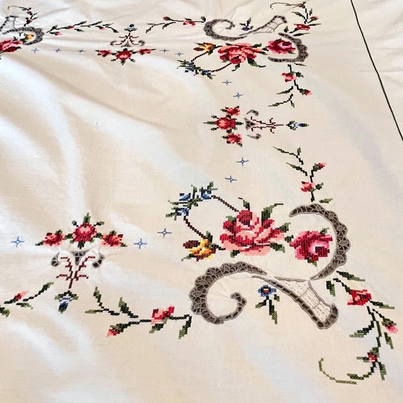 May include: White tablecloth with intricate cross-stitch floral embroidery. The design features red, pink, and blue flowers with green leaves and stems. Decorative gray lace-like accents frame the floral patterns.