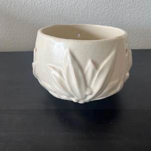 Op de afbeelding: Een crèmekleurige keramische kom met een ronde vorm en een brede opening. Het buitenoppervlak is voorzien van een verhoogd, reliëf bladpatroon dat rondom het lichaam loopt. Kleine ronde gaatjes zijn zichtbaar nabij de bovenste rand.