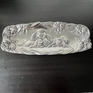 May include: A silver-colored rectangular tray with a raised relief design of a family of rabbits surrounded by flowers. The tray has a smooth, reflective surface and is likely made of metal.