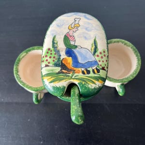 May include: A vintage ceramic sugar bowl with a lid featuring a hand-painted scene of a woman in traditional attire. The bowl has a green trim and a matching spoon. The interior is cream-colored.