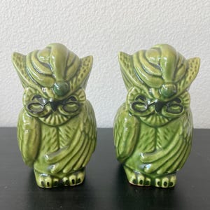 May include: Two ceramic owl figurines in a glossy green glaze. The owls have closed eyes, detailed feather patterns, and small feet. The figurines are displayed on a dark surface.