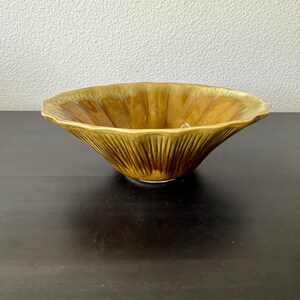 Könnte beinhalten: Eine breite, flache Keramikschale mit einer glänzenden, senfgelben Glasur und dunkelbraunen vertikalen Streifen, die von der Mitte zum gewellten, geschwungenen Rand verlaufen. Die Schale hat eine konische Form mit einer glatten Oberfläche.