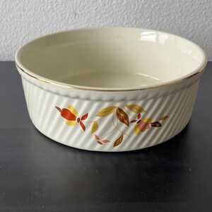 May include: A round, cream-colored ceramic bowl featuring a diagonal ribbed texture on the exterior. The rim is accented with a thin gold band. A floral pattern in shades of orange, yellow, and brown is printed on the front of the bowl.