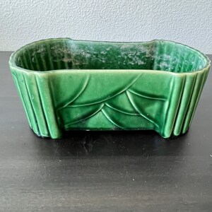 May include: A green ceramic planter with a rectangular shape and a glossy finish. The planter features a leaf design on the sides and a ribbed texture. This decorative piece is suitable for indoor or outdoor use.