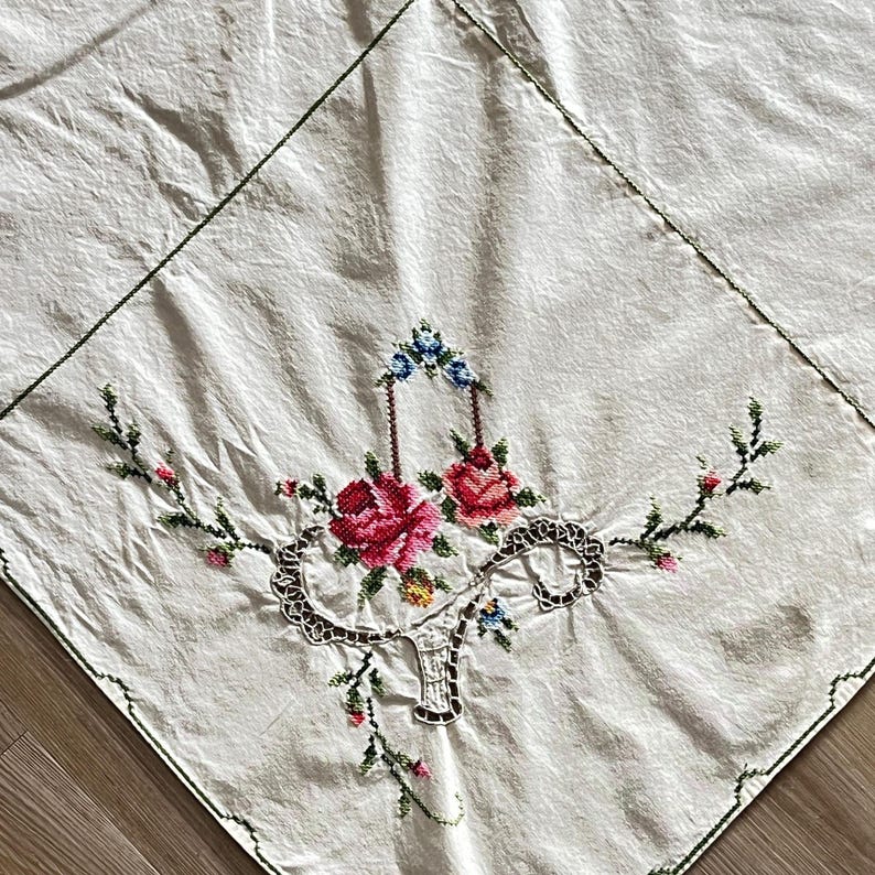 May include: Cream-colored square tablecloth with a floral embroidery design. The embroidery features pink and red roses, blue and yellow accents, and green stems. The edges are outlined with green stitching.