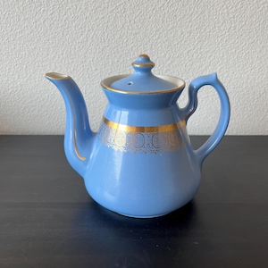 Op de afbeelding: Een lichtblauwe keramische theepot met een gouden band met een sierlijk ontwerp. De theepot heeft een gebogen tuit, een handvat en een deksel met een kleine knop. De rand van het deksel is wit. De theepot staat op een donkere ondergrond.