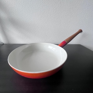 Könnte beinhalten: Eine rot-weiße Bratpfanne mit Holzgriff. Die Pfanne hat eine weiße Innenseite und eine leuchtend rote Außenseite. Der Griff ist hellbraun mit einem roten Akzent in der Nähe der Pfanne.