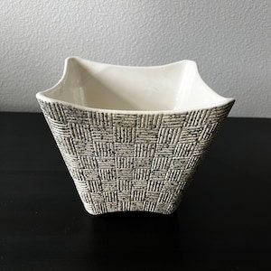 May include: A square ceramic bowl with a flared, wavy rim. The exterior features a textured, grey and white basket-weave pattern with a speckled finish, while the interior is smooth, solid white. The bowl has a wide opening and a tapered base.