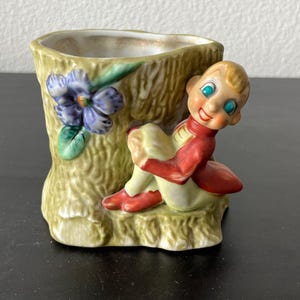 May include: A vintage ceramic planter shaped like a tree trunk, with a smiling elf figurine. The planter is painted in shades of green, with a blue flower and the elf wearing a red jacket and shoes.