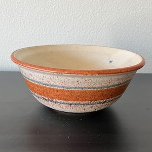 Può includere: Una ciotola in ceramica con esterno beige maculato e bordo marrone rossiccio. La ciotola presenta due fasce orizzontali di strisce arancioni e grigie. L'interno è di colore beige chiaro.