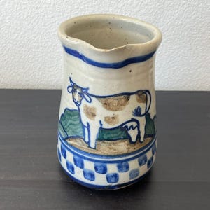 May include: A ceramic pitcher with a cream-colored base, featuring a hand-painted brown and white cow standing in a green landscape. The rim and base are decorated with blue accents, including a blue checkered pattern around the bottom edge.