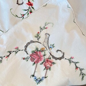 May include: Close-up of a white embroidered tablecloth with floral and vase designs. The embroidery features pink, red, and blue flowers with green leaves. The vase design is gray with a decorative border.