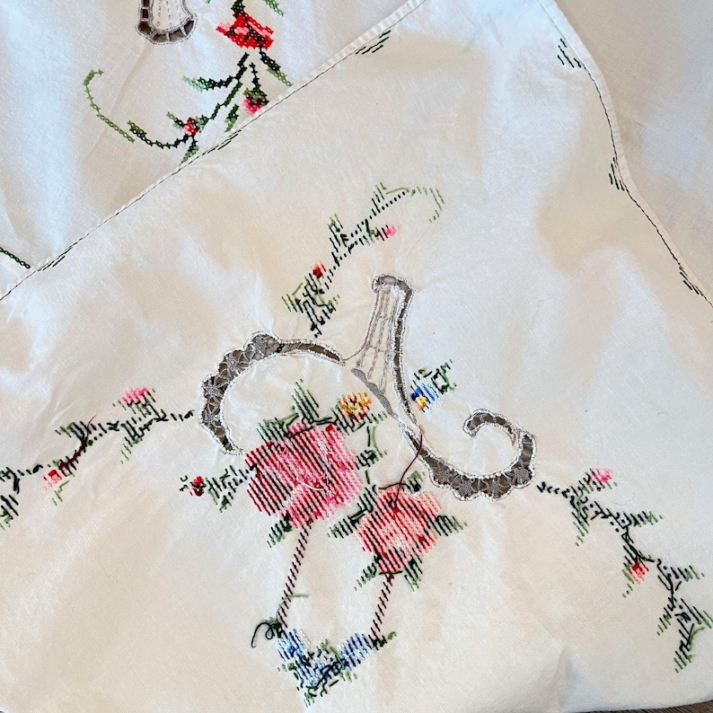 May include: Close-up of a white embroidered tablecloth with floral and vase designs. The embroidery features pink, red, and blue flowers with green leaves. The vase design is gray with a decorative border.