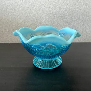 May include: A vintage, light blue glass bowl with a scalloped rim and a pedestal base. The bowl features a detailed grape and leaf pattern. The glass has a slight iridescence, adding to its antique charm. A decorative piece for serving or display.