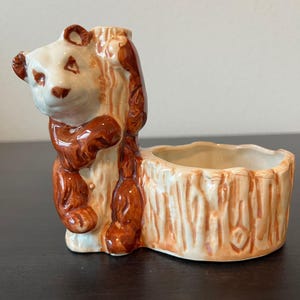 Puede incluir: Una maceta de cerámica con forma de oso panda en marrón y crema. El panda se aferra a una maceta con forma de tronco de árbol. La maceta tiene una abertura redonda para plantas u otros objetos decorativos. La cara del panda es de color crema con detalles marrones.