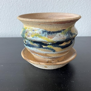 May include: A handcrafted ceramic planter with a matching saucer. The planter features a layered design with bands of tan, blue, yellow, and black glaze. The rim and saucer have a natural, unglazed appearance.