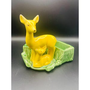 Pode incluir: Uma estatueta de veado em cerâmica amarelo-brilhante ao lado de um filhote menor, ambos fixados numa base de vaso retangular verde-claro com textura moldada de relva e tronco. O veado tem olhos e orelhas pintados de castanho e o vaso é oco.