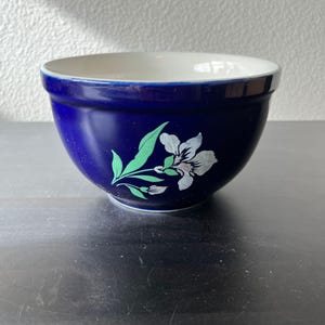 May include: A deep blue ceramic mixing bowl with a smooth, glossy finish. The exterior features a decorative floral motif consisting of a single white flower with green leaves and a stem. The interior of the bowl is a solid, clean off-white colour.