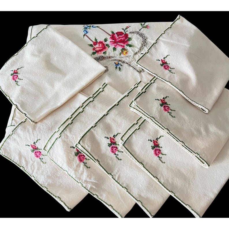 May include: A cream-colored embroidered tablecloth and matching napkins. The tablecloth features a floral design with red and pink roses, green leaves, and blue accents. The napkins have a smaller floral embroidery and green trim.